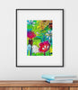 Framed colorful abstract art piece on a white wall with books on a wooden surface below.