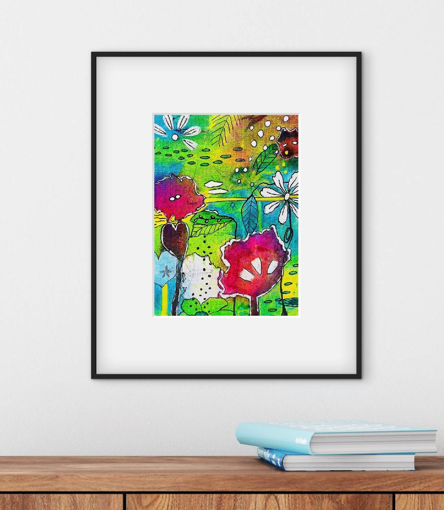 Framed colorful abstract art piece on a white wall with books on a wooden surface below.