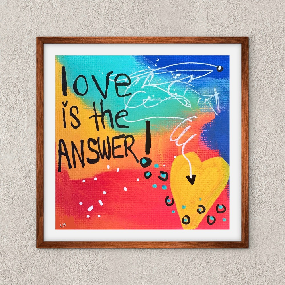 Framed abstract art print on a textured light gray wall. The artwork features the text 'love is the ANSWER!' in black over a colorful gradient background of blue, yellow, orange, and red, with a large yellow heart and white scribbled lines.