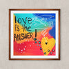 Framed abstract art print on a textured light gray wall. The artwork features the text 'love is the ANSWER!' in black over a colorful gradient background of blue, yellow, orange, and red, with a large yellow heart and white scribbled lines.