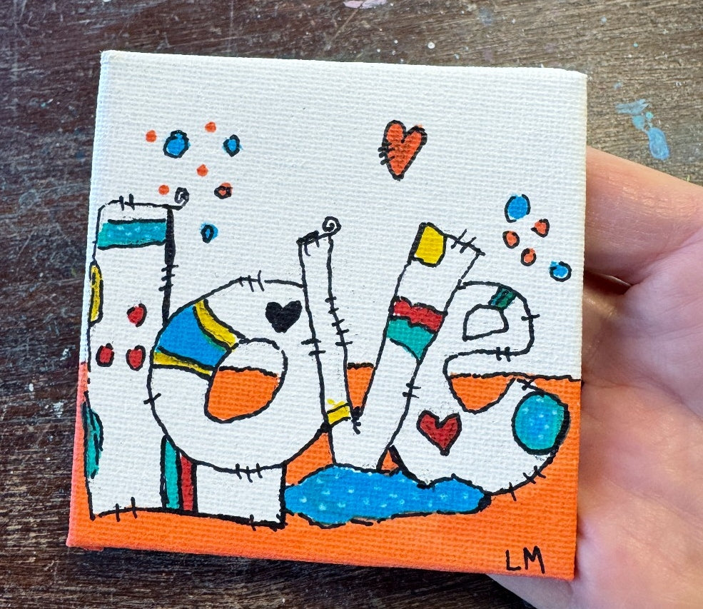 A hand holding a small square canvas painting that spells out 'LOVE' in a colorful, whimsical patchwork style, set against a dark wooden background.