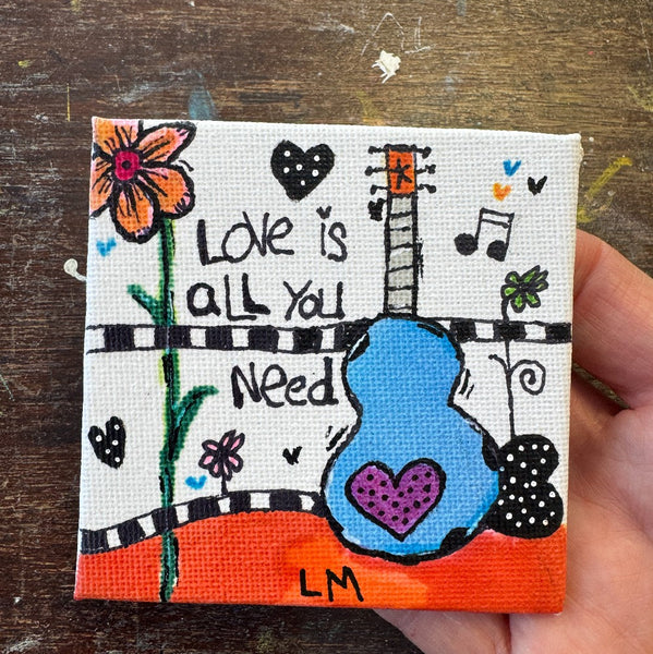 A small square canvas painting held by a hand, featuring a colorful hand-drawn guitar, flowers, hearts, and the text 'Love is all you need' on a rustic wooden background.