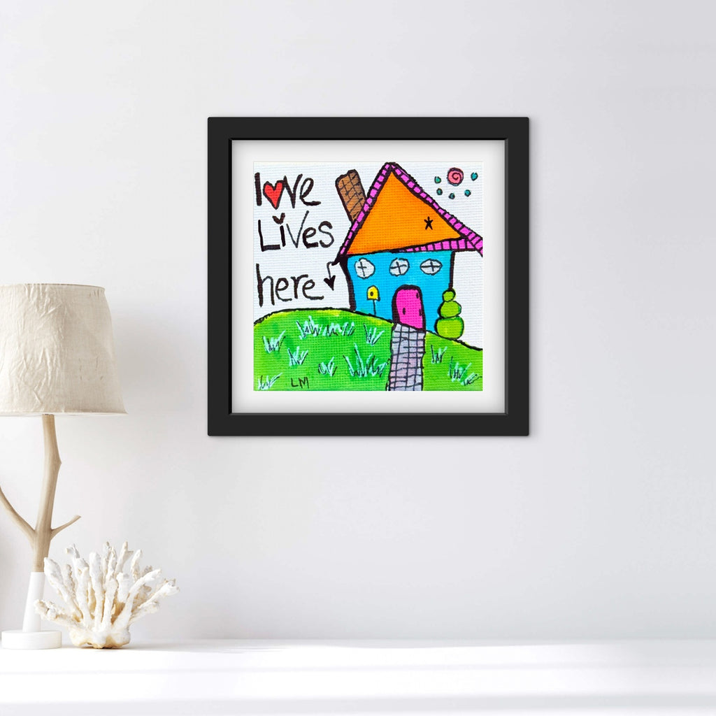 Framed artwork with a colorful house and 'Love Lives Here' text on a white wall.