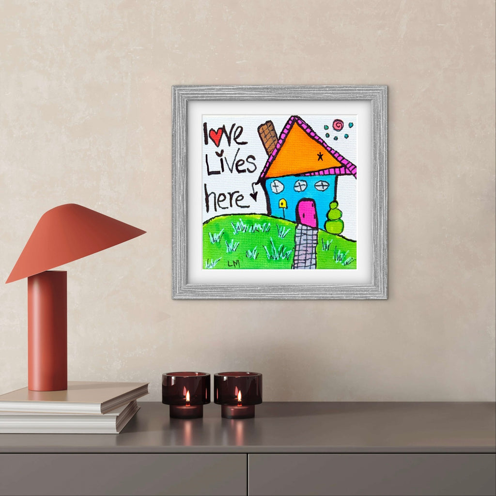 Framed children's drawing of a house with 'Love Lives Here' text on a wall above a desk.