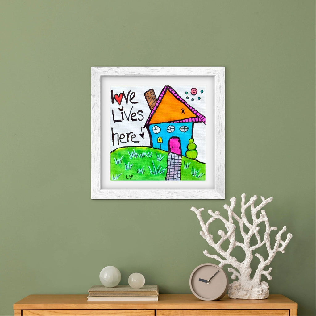 Framed children's drawing of a house with 'Love Lives Here' text on a green wall.