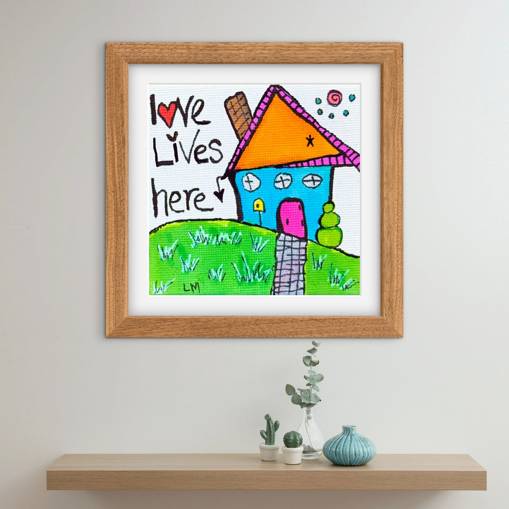 Framed children's drawing of a house with 'Love Lives Here' text on a wall above a shelf.