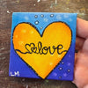 A small square canvas painting held by a hand, featuring a large yellow heart with the word 'love' in black script over a blue and purple background with small white dots.