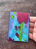 A small abstract floral painting held by a hand over a scratched dark wood surface. The artwork features a bright blue background with a large magenta flower, green stem and leaves, and smaller dark pink blossoms.