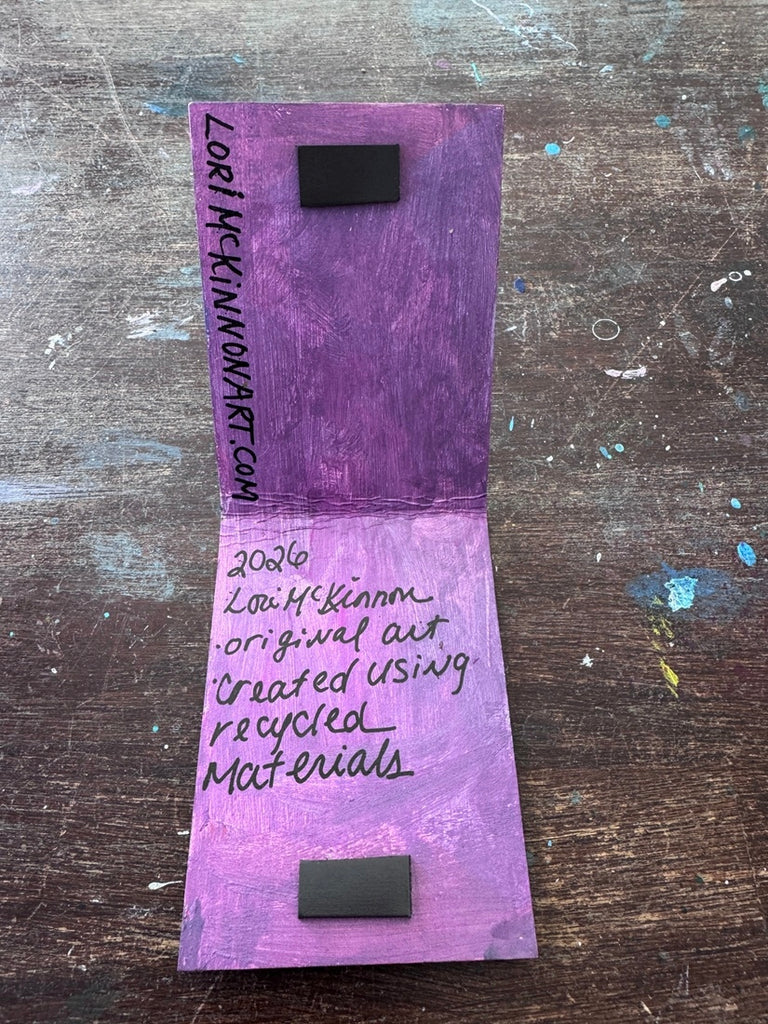 A handmade purple and lavender paper art piece with black magnetic closures, featuring handwritten text that reads 'original art Created using recycled Materials' displayed on a rustic wooden background.