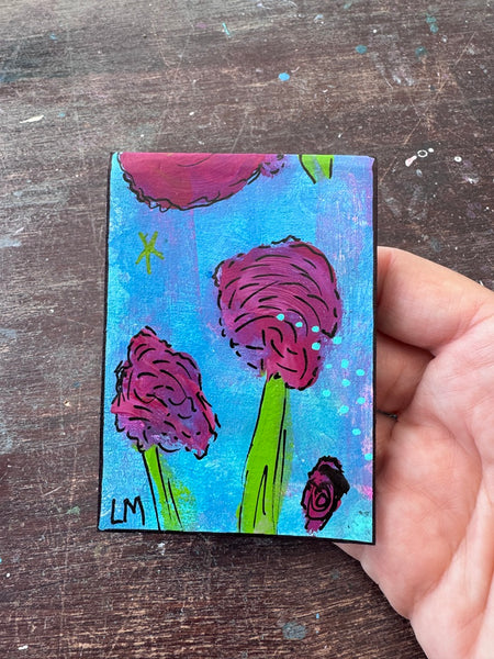 A hand holding a small painting with a colorful abstract floral design, set against a dark wooden background.
