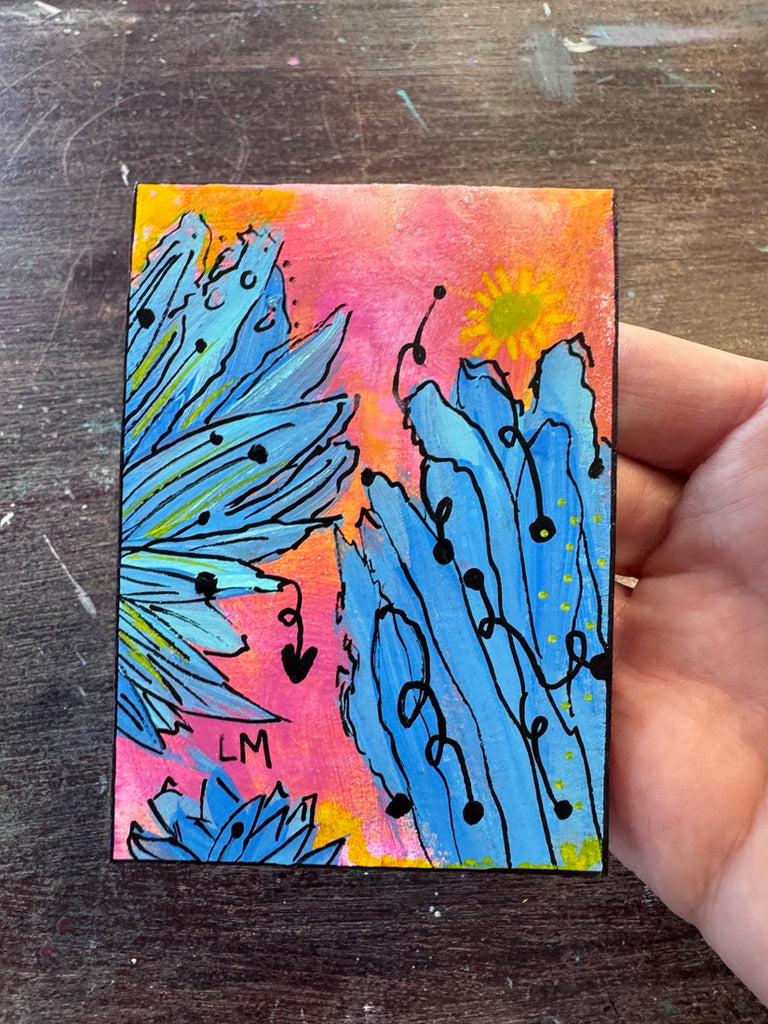 A hand holding a small abstract painting with blue floral-like forms, pink and orange gradients, black line details, and a yellow sun on a dark wooden surface.