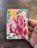 A hand holding a small art print or painting featuring pink flowers with teal leaves on a peach and yellow background, set against a dark wooden surface.