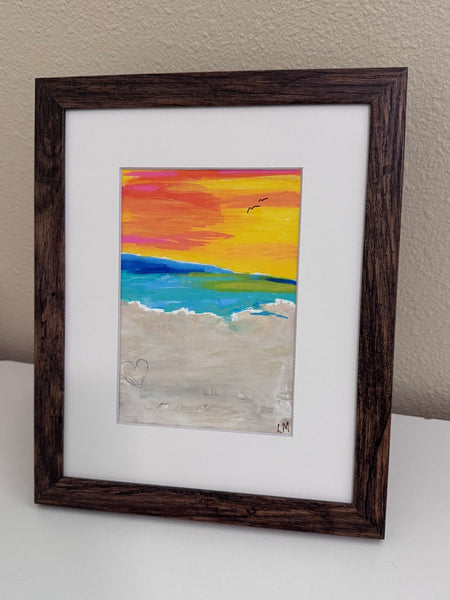A framed colorful beach sunset painting with a dark brown wooden frame and white mat, leaning against a beige wall on a white surface.