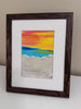 A framed colorful beach sunset painting with a dark brown wooden frame and white mat, leaning against a beige wall on a white surface.