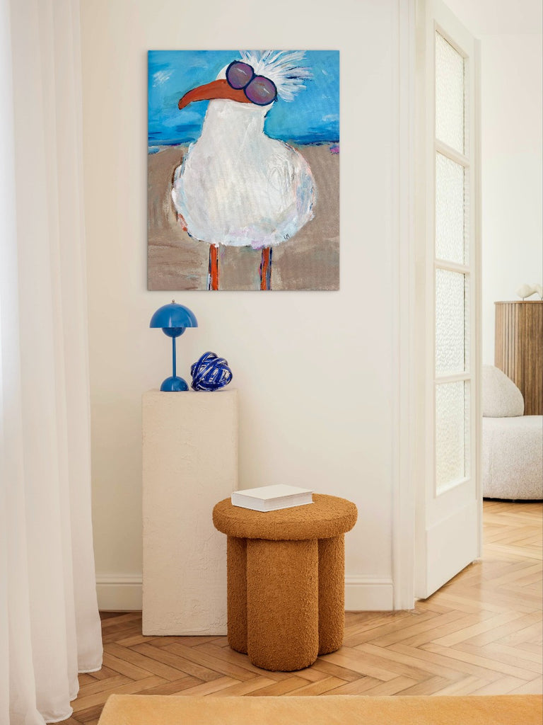 Painting of a bird with sunglasses on a wall above a white pedestal with decorative items.