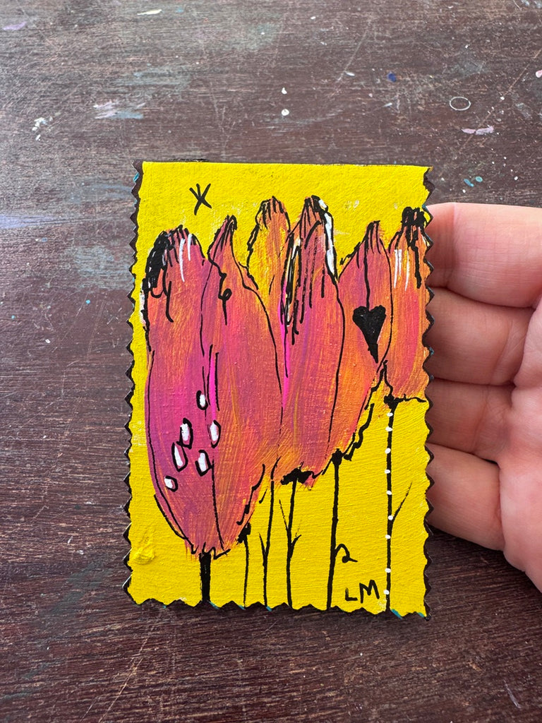 A hand holding a small painting featuring a floral painting in pink, orange, and yellow on a dark wooden surface.