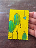 A hand holding a small yellow bookmark with a whimsical painting of green and blue hanging pods on black stems, set against a dark wooden tabletop.