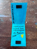 Inside of a handmade blue bookmark with jagged edges, featuring black magnetic strips at the top and bottom, handwritten text in black ink, and a small yellow heart. The card is photographed flat on a worn, paint-splattered wooden table.