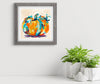 Framed abstract art of a pumpkin on a white wall with plants below.
