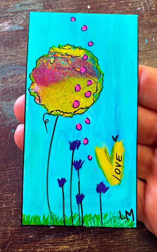 A hand holding a small painted  bookmark featuring a bright turquoise background, a large yellow flower with pink dots, purple flowers, green grass, and a yellow heart with the word LOVE.