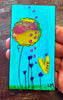 A hand holding a small painted  bookmark featuring a bright turquoise background, a large yellow flower with pink dots, purple flowers, green grass, and a yellow heart with the word LOVE.
