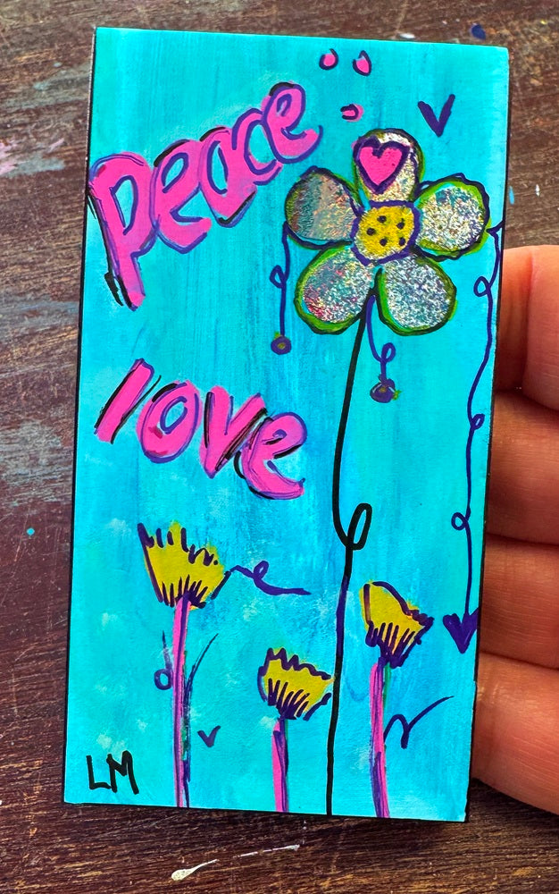 A small painted bookmark held in a hand, featuring the words 'Peace love' in pink over a bright turquoise background, with a flower and smaller yellow flowers. The hand is visible on the right side over a dark wooden surface