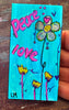 A small painted bookmark held in a hand, featuring the words 'Peace love' in pink over a bright turquoise background, with a flower and smaller yellow flowers. The hand is visible on the right side over a dark wooden surface