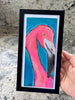 a hand holding a flamingo painting in a black frame over a quartz counter