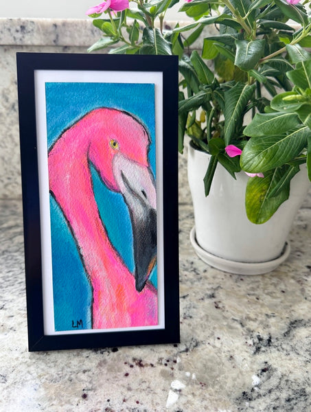 a framed flamingo painting standing on a counter next to a plant with pink flowers