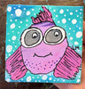 A square canvas painting of a whimsical purple and pink fish with large gray eyes, set against a turquoise background with white polka dots. The canvas is held by a hand over a dark wooden surface.