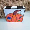 pumpkin art on a wood block sitting on a white table in front of a basket