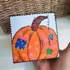 pumpkin art on a wood block being held by a hand over a white table in front of a basket