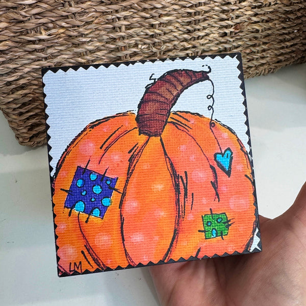 pumpkin art on a wood block being held by a hand over a white table in front of a basket