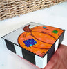 pumpkin art on a wood block being held by a hand over a white table in front of a basket