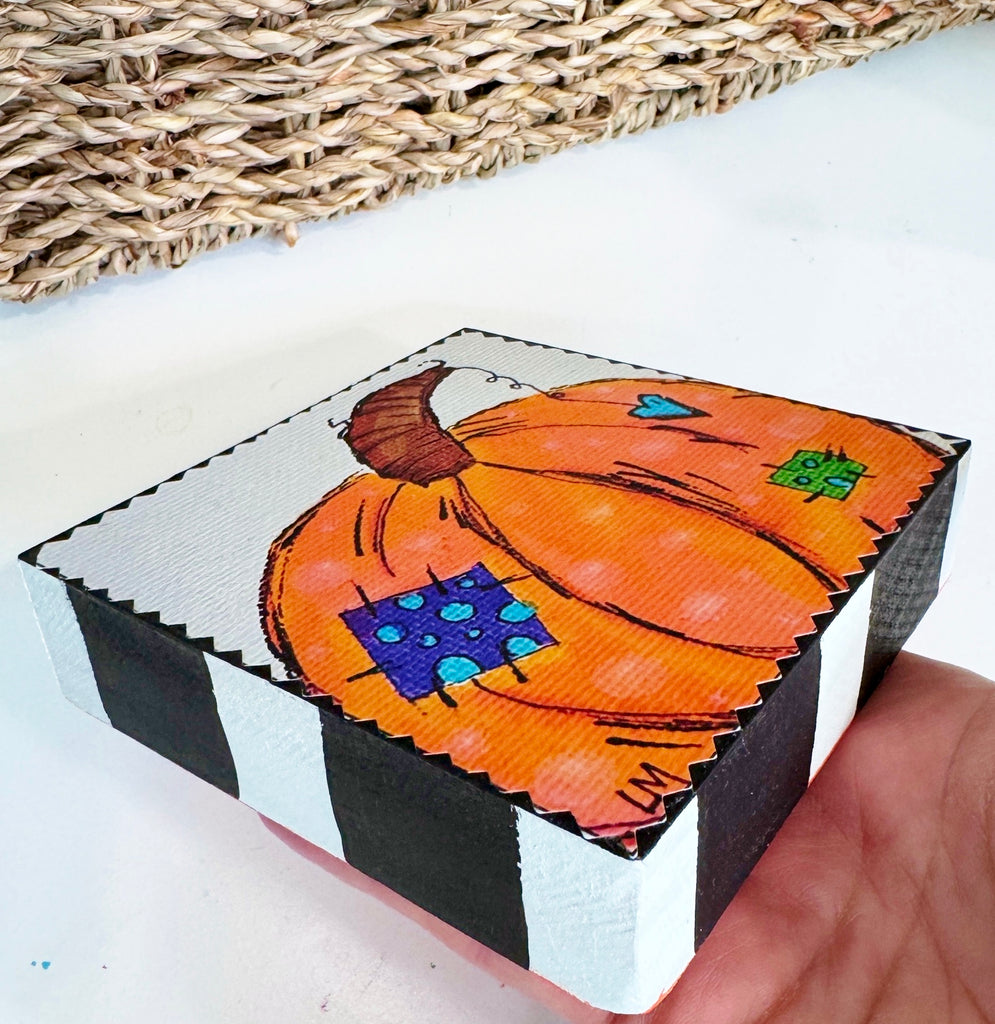 pumpkin art on a wood block being held by a hand over a white table in front of a basket