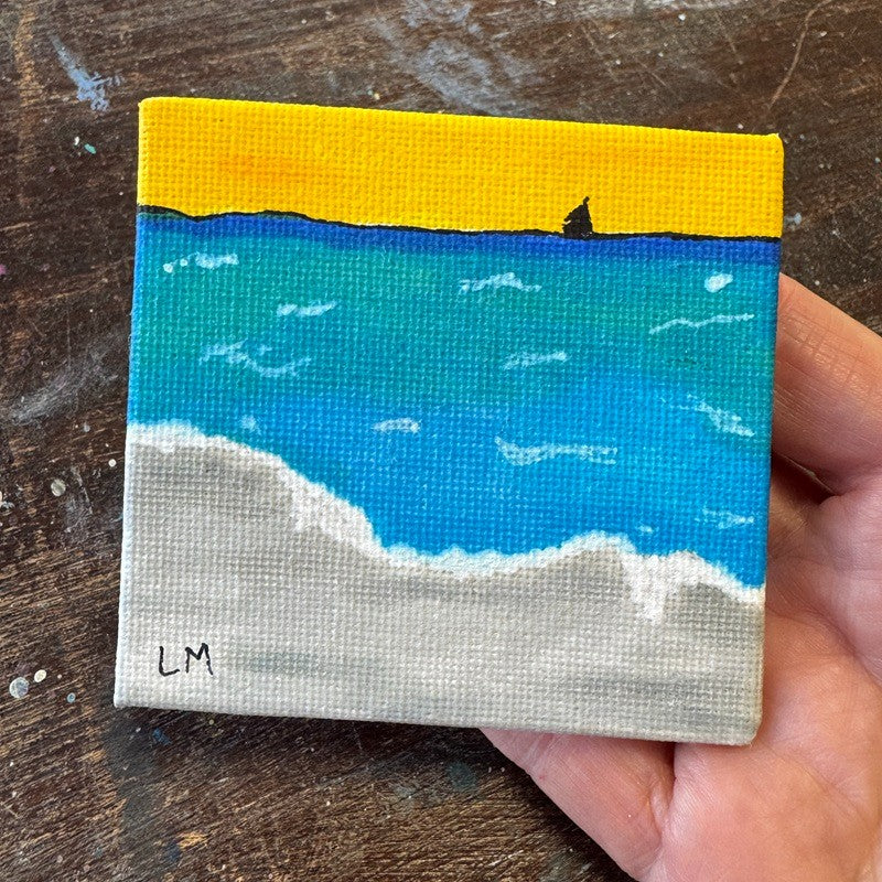 A square canvas painting of a beach scene with a yellow sky, blue ocean, white waves, and a light gray shore, held in a person's hand over a dark wooden surface.