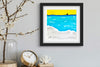 Framed artwork of a beach scene with a yellow sky, blue water, and white clouds on a gray wall.