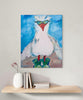 A vertical canvas art print featuring a whimsical painting of a white bird with green sunglasses and a red beak, displayed on a light gray wall above a wooden shelf with books and a white vase.