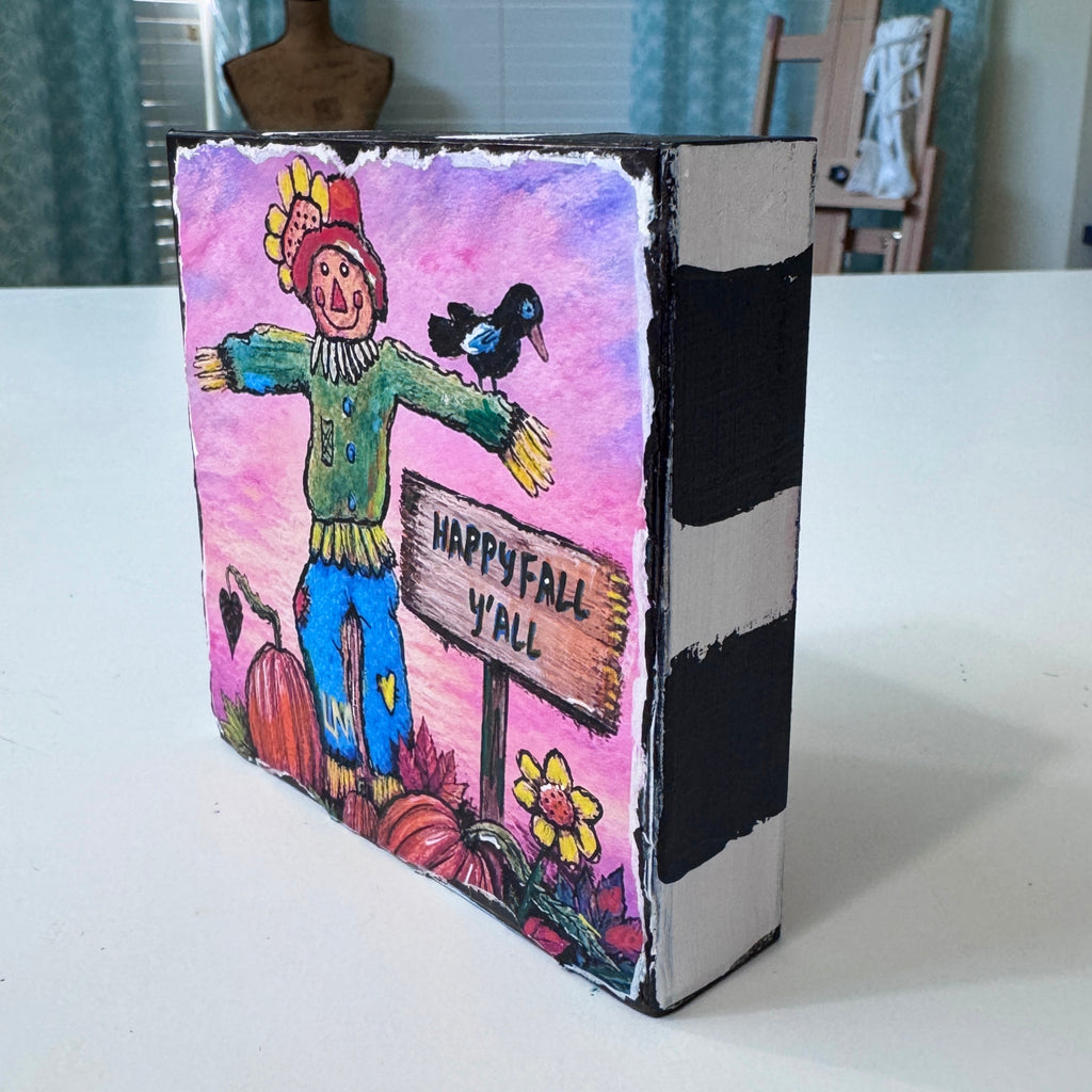 whimsical scarecrow artwork with a sign that says "happy Fall Y'all". Scarecrow is in a pumpkin patch with a crow, pumpkins and flowers