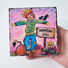whimsical scarecrow artwork held by a hand. A sign on the art says "happy Fall Y'all". Scarecrow is in a pumpkin patch with a crow, pumpkins and flowers