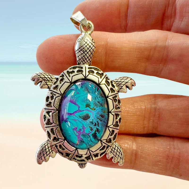 Tortoise-shaped pendant with turquoise stone held between fingers against a beach background
