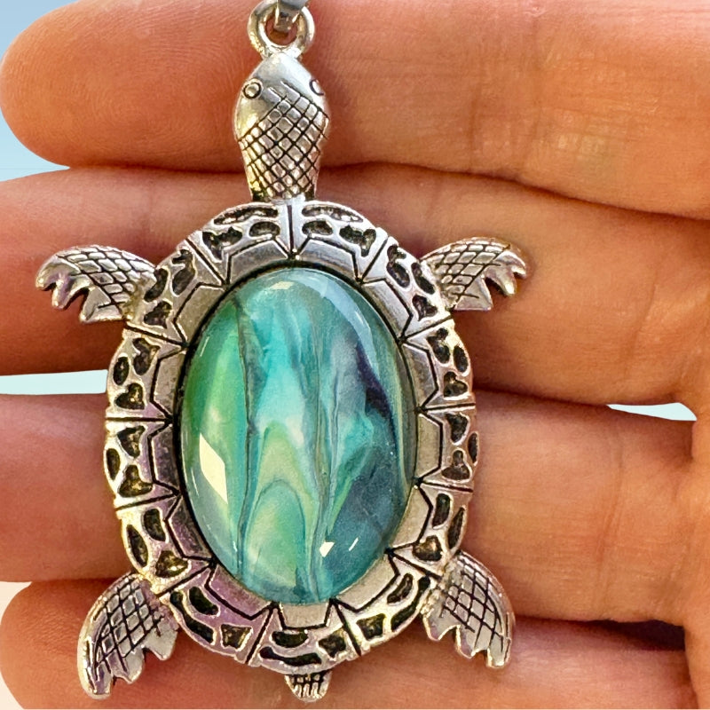 Turtle shaped pendant with a blue art glass in the center being held by a hand