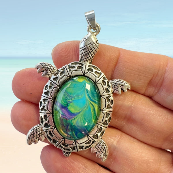 turtle pendant with a painted glass center held between fingers against a beach background
