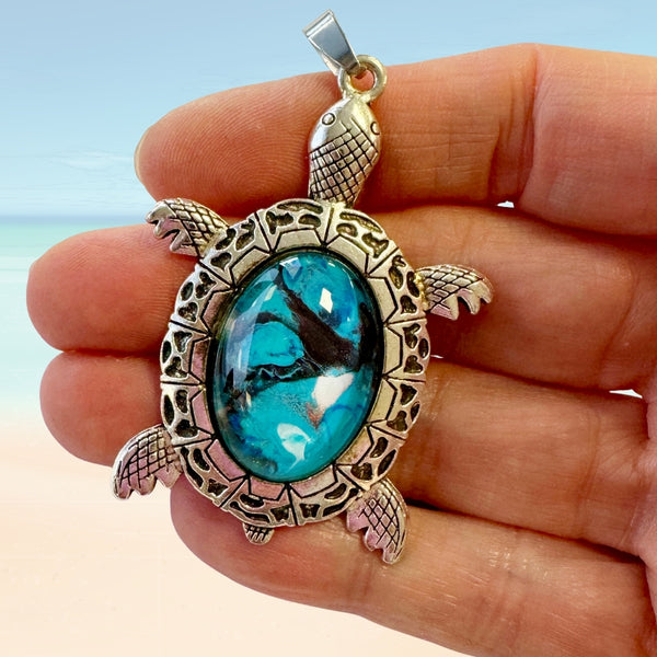 Turtle shaped pendant with glass center held in a hand against a beach background