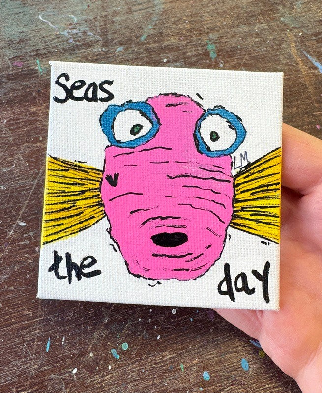 A hand holding a small square canvas painting with a whimsical pink fish face, blue eyes, yellow rays, and the handwritten text 'Seas the day'. The canvas is photographed over a worn wooden surface.