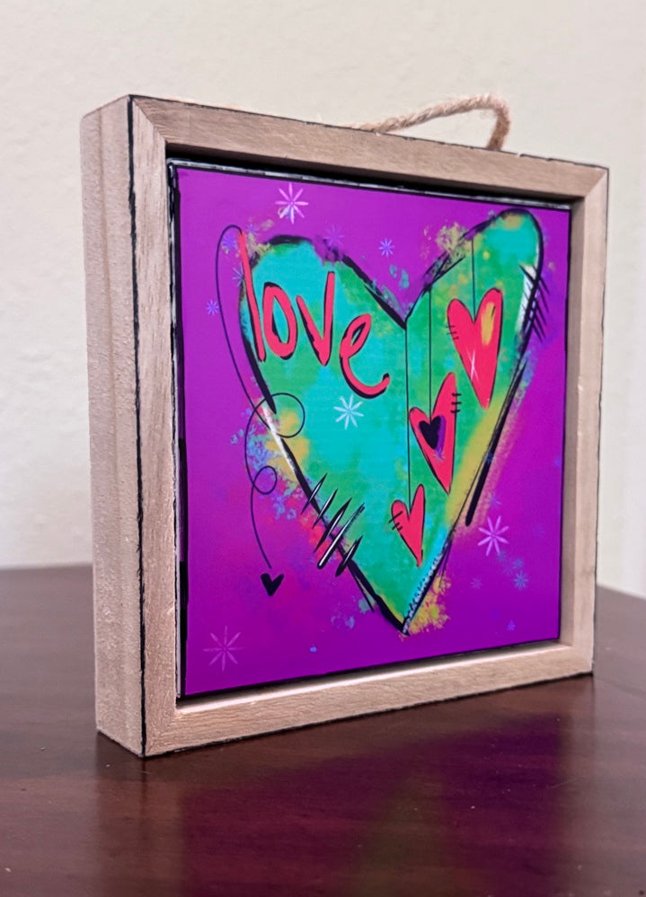A square framed art print featuring a colorful heart design on a purple background, displayed on a dark wooden surface against a light wall.