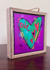 A square framed art print featuring a colorful heart design on a purple background, displayed on a dark wooden surface against a light wall.