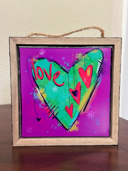 A square framed art print with a rustic wooden frame and hanging rope, featuring a colorful heart design on a purple background with the word 'love' in red script.