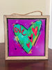 A square framed art print with a rustic wooden frame and hanging rope, featuring a colorful heart design on a purple background with the word 'love' in red script.