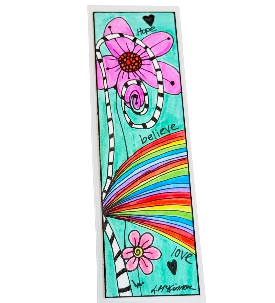 Laminated bookmark featuring a colorful hand-drawn design with pink flowers, a rainbow, black hearts, and the words 'hope', 'believe', and 'love' on a turquoise background. Signed by artist in lower right 